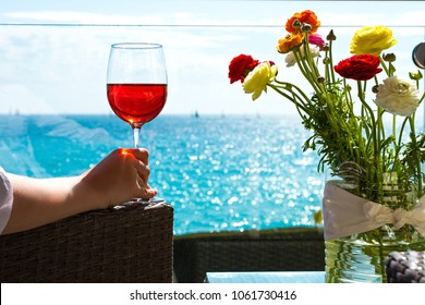 Elegant female hand with a glass of pink wine and flowers against the background of a view of the sea.