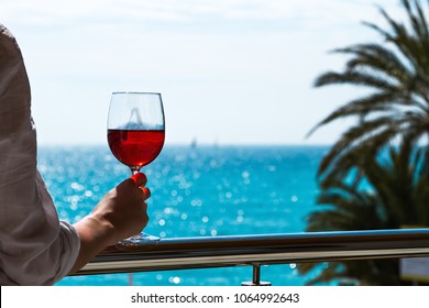 The elegant female hand holding a glass of red wine with the background of a sunny seaside view.