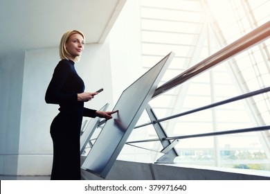 Elegant Female Holding Mobile Phone While Standing Front High Tech Digital Device In Office Interior, Young Female Thinking While Searching Information On Cell Telephone And Interactive Display