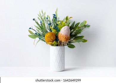 A Elegant Floral Arrangement In A White Vase On A White Table With A White Background. Flower Bunch Includes Blue And Yellow Flowers, Pink And Yellow Australian Native Banksias And Eucalyptus Leaves.