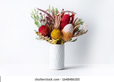 Elegant Flower Arrangement Of Mostly Australian Native Dried Banksias, Eucalyptus Leaves And Gum Nuts, Fountain Grass And Dried Red Flowers, In A White Vase On A Table With A White Background.