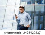 Elegant gentleman in a blue shirt, smiling while using a smartphone and holding a laptop, near a modern glass building.

