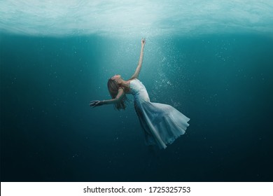 Elegant Girl Dancer In White Dress In A State Of Levitation Under The Deep Waters Of The Ocean With Sunlight Beaming On Her Face. 