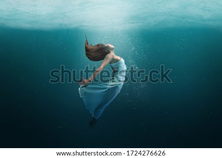 Elegant girl dancer in white dress in a state of levitation under the deep waters of the ocean with sunlight beaming on her face. 