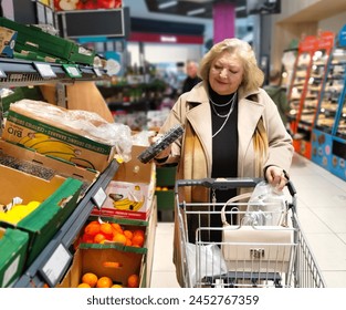 Elegant, good looking senior woman doing shopping in a supermarket. Mature woman with a cart filled with groceries in front of the shelves with fruit and vegetables. - Powered by Shutterstock