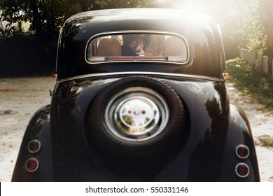 elegant gorgeous bride and handsome groom embracing in stylish black car in light. unusual view from back. luxury wedding couple in retro style.