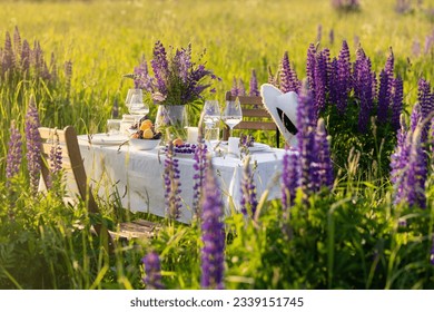 Elegant gorgeous wedding table decor or romantic dinner arrangement outdoors in blooming field. Purple lupine flowers, candles, fruits and wine, wooden vintage furniture. Sunset, summer, golden hour