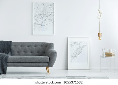 Elegant, gray sofa with wooden legs and large map posters on a white wall in a designer minimalist living room interior of an architect