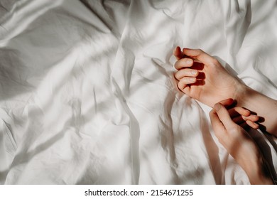 Elegant hands lie on the white bed sheet in the sunlight. Bed with white linens. The concept of a good morning, stress relief, self-care, relaxation and time for yourself