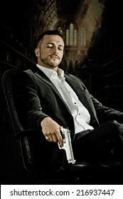 Elegant Handsome Latin Man Gangster Mafia Spy Hitman Assassin With Closed Eyes Sitting In A Chair Holding A Gun Over Dark Background