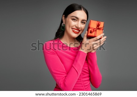elegant and happy woman with earrings and brunette hair smiling while holding red and wrapped present for holiday on grey background