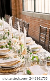 An elegant head table in a brick warehouse. The table is decorated with candlesticks, plates and menus, and greenery and florals.
