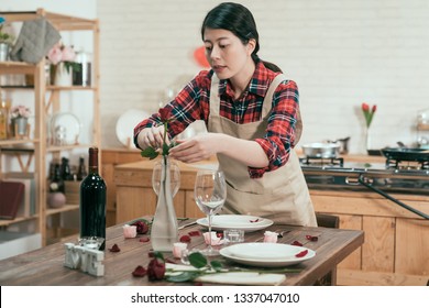 elegant housewife in apron decorated table prepared for valentine day dinner at home. young asian woman in pinafore set up table putting flower in vase in middle on desk next red wine bottle glasses - Shutterstock ID 1337047010