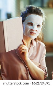 elegant housewife in pajamas with white sheet facial mask looking out from book at modern home in sunny winter day.