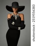 Elegant Lady in Black Dress wearing Pearl Jewelry Necklace and Wide Brim Hat. Fashion Woman in Cocktail Form Fitted Gown and Gloves with Bracelets over Gray. Stylish Model with hidden Face