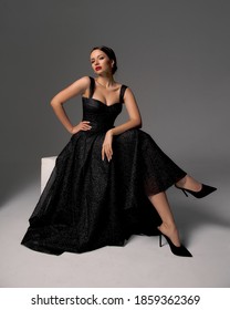 Elegant Lady In Black Shiny Ball Gown And Shoes Sitting At White Cibe With Crossed Legs And Posing Against Grey Background, Full Length Fashion Portrait. Sexy Female Model