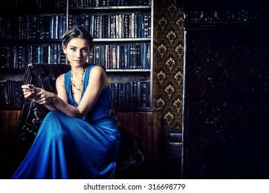 Elegant lady wearing evening dress sitting in the chair in the old vintage library. Beauty, fashion.