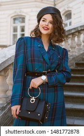 Elegant lady wearing trendy autumn blue checkered dress, leather beret, wrist watch, holding black bag, posing in street of European city