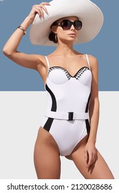 Elegant lady in white wide-brimmed hat and sunglasses is wearing black and white one-piece swimsuit with belt. Lady is posing on blue and white background and holding brim of her hat.