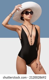 Elegant lady in white wide-brimmed hat and sunglasses is wearing black one-piece swimsuit with deep neck, straps and rings. Lady is posing on blue and white background and holding brim of her hat.