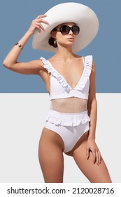 Elegant lady in white wide-brimmed hat and sunglasses is wearing white one-piece swimsuit with mesh and ruffles. Lady is posing on blue and white background and holding brim of her hat.