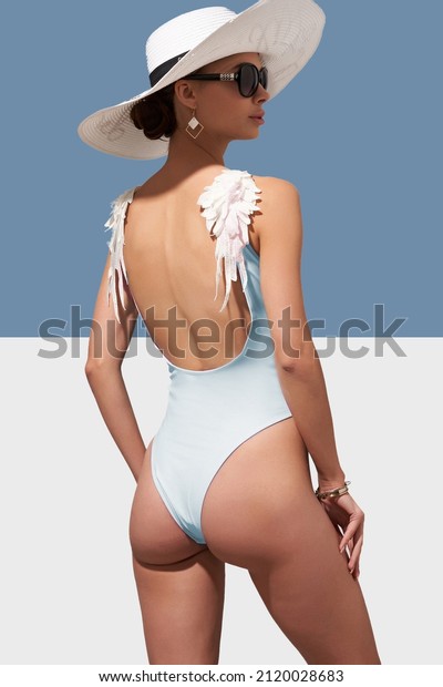 Elegant lady in\
white wide-brimmed hat and sunglasses is showing backside of pale\
blue one-piece swimsuit with wings on shoulders. Lady is posing on\
blue and white\
background.
