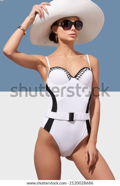 Elegant\
lady in white wide-brimmed hat and sunglasses is wearing black and\
white one-piece swimsuit with belt. Lady is posing on blue and\
white background and holding brim of her\
hat.
