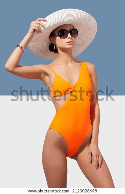 Elegant lady in\
white wide-brimmed hat and sunglasses is wearing orange asymmetric\
one-piece swimsuit. Lady is posing on blue and white background and\
holding brim of her\
hat.