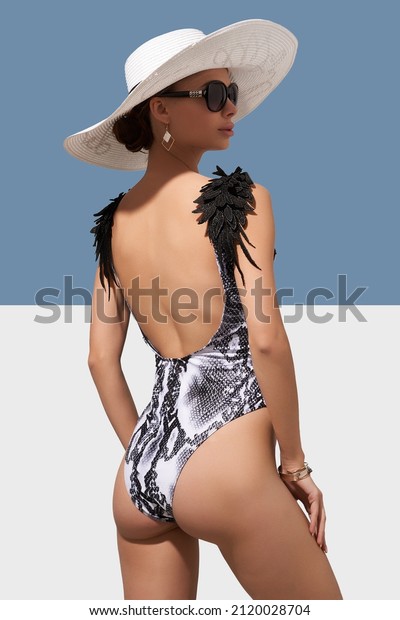 Elegant lady in white wide-brimmed hat and
sunglasses is showing backside of black and white one-piece
swimsuit with wings on shoulders and snake print. Lady is posing on
blue and white
background.