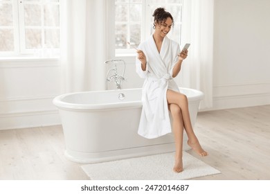 Elegant Latin woman in white robe enjoying her morning routine while sitting on the edge of bathtub, interacting with her smartphone and holding toothbrush - Powered by Shutterstock