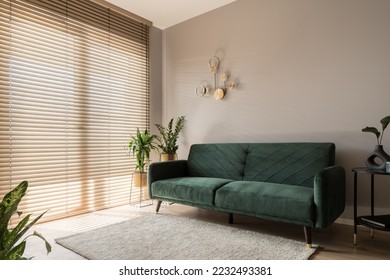 Elegant living room with big window wall behind wooden blinds, comfortable green sofa, carpet and decorative gold wall lamp - Shutterstock ID 2232493381