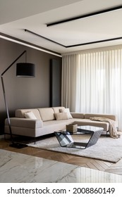 Elegant living room with stylish, beige, corner sofa, modern coffee table, simple black lamp and big windows behind curtains
