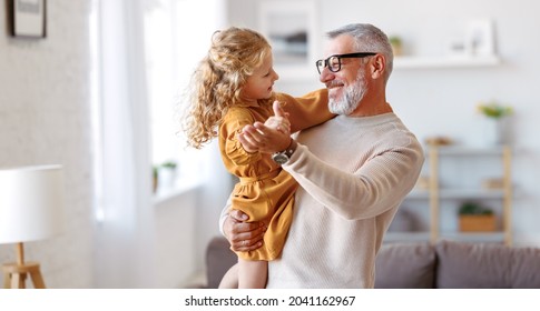 Eleganter liebevoller, fürsorglicher Großvater, der seine süße kleine Enkeltochter, das bezaubernde Mädchen und das positive Großvater beim Tanzen zusammen im Wohnzimmer zu Hause ansieht. Familienbegriff