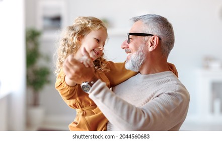 Elegant loving caring grandfather looking at his cute little granddaughter, adorable child girl and positive grandpa holding hands while dancing together in living room at home. Family concept - Shutterstock ID 2044560683
