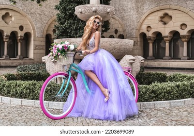 Elegant Luxury Fashion. Glamour, Stylish Elegant Woman In Amazing Long Gown Dress On Colorful Bike. Female Model In Lilac Long Dress In The City. Travel Model. Outdoor Shoot. Vogue. Couture.