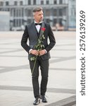 elegant man in black tux. man wearing tux bowtie outdoor. tux man with red rose walking outdoor