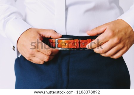 An elegant man in blue pants and a white formal shirt dresses a brown leather trouser belt. 
