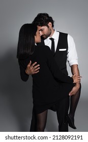 Elegant Man Embracing Passionate Asian Woman In Black Blazer Dress On Grey Background