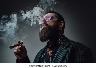 Elegant man in retro style poses in dark background smoking cigar. Portrait of a bearded guy with stylish coiffure.