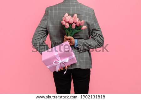 Elegant man in suit holding a festive pink box and a bouquet of flowers behind his back on a pink background, a gift for womens day, Valentine's Day concept