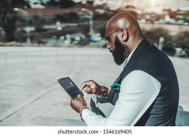 An elegant mature bald African man with a beard and in spectacles is using outdoors his futuristic gadget with a user interface and infographics on it to implement a secure login using his fingerprint