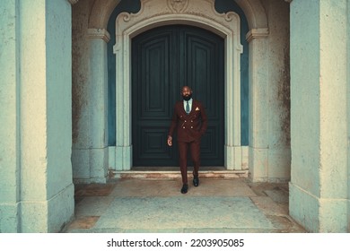 An Elegant Mature Bald Bearded African Man In A Costume And Glasses Is Leaving A Wooden Entry Gate Of An Antique Building; A Dapper Black Guy In A Tailored Suit Walked Out The Door Of An Old House