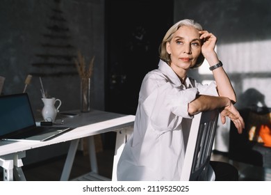 Elegant Mature Caucasian Female Entrepreneur Sitting On Chair And Looking Away. Concept Of Small Business And Entrepreneurship. Modern Successful Woman At Dark Art Studio With Sunny Lighting