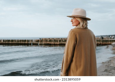 Elegant mature lady in coat and round brown hat standing on seashore outdoors, back view of lonely senior woman enjoying sea looking at horizon. - Powered by Shutterstock