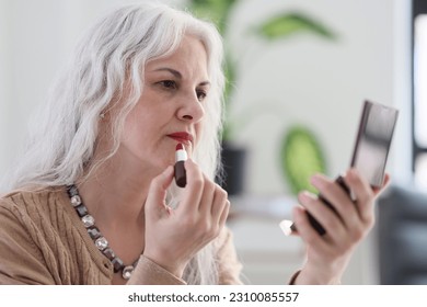 Elegant mature woman with long grey hair applies red lipstick looking at mirror. Attractive senior lady applies makeup sitting in light room - Powered by Shutterstock