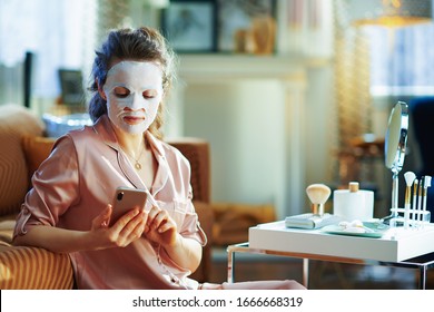 Elegant Middle Age Woman In Pajamas With White Sheet Facial Mask On Face Cloud Download To Smartphone From Stored Data On Server In The Modern Living Room In Sunny Winter Day.