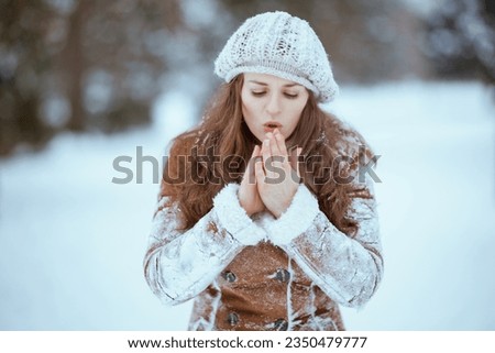 elegant middle aged woman in brown hat and scarf in sheepskin coat warming cold hands with breath outside in the city in winter.