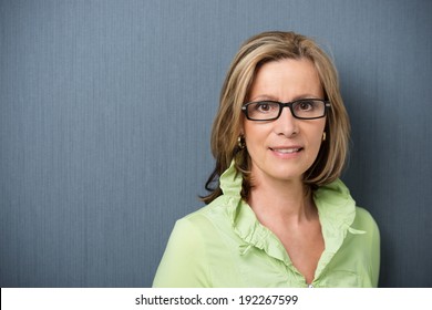 Elegant middle-aged woman in glasses looking directly at the camera with a friendly smile and interested expression, on grey with copyspace