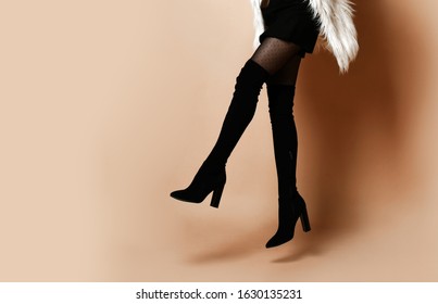 Elegant model with long hair, in brown turtleneck, faux fur sleeveless coat, short black skirt, over the knee boots, bracelet. Smiling, jumping up, posing on beige background. Full length, copy space