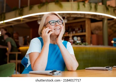 Elegant Modern Older Mature Senior Woman With Glasses Is Sitting In A Cafe With A Mobile Phone With A Cup Of Tea. The Retirement Age Is 60 Years.